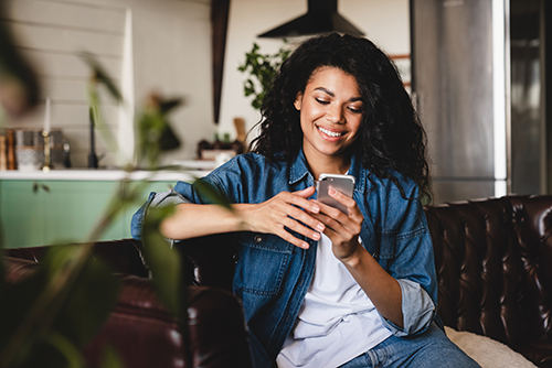 Woman sitting on couch smiling at phone next to a plant.